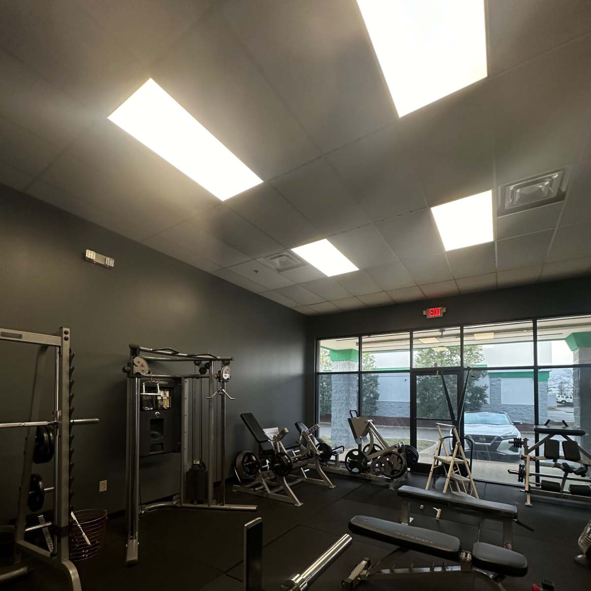 A fitness room with various exercise equipment, including weight benches, a cable machine, and stationary bikes, all arranged along the perimeter near large windows.