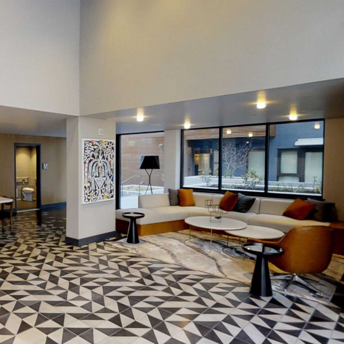 A modern lobby with geometric tile flooring, a seating area by large windows, and a long table with hanging pendant lights.