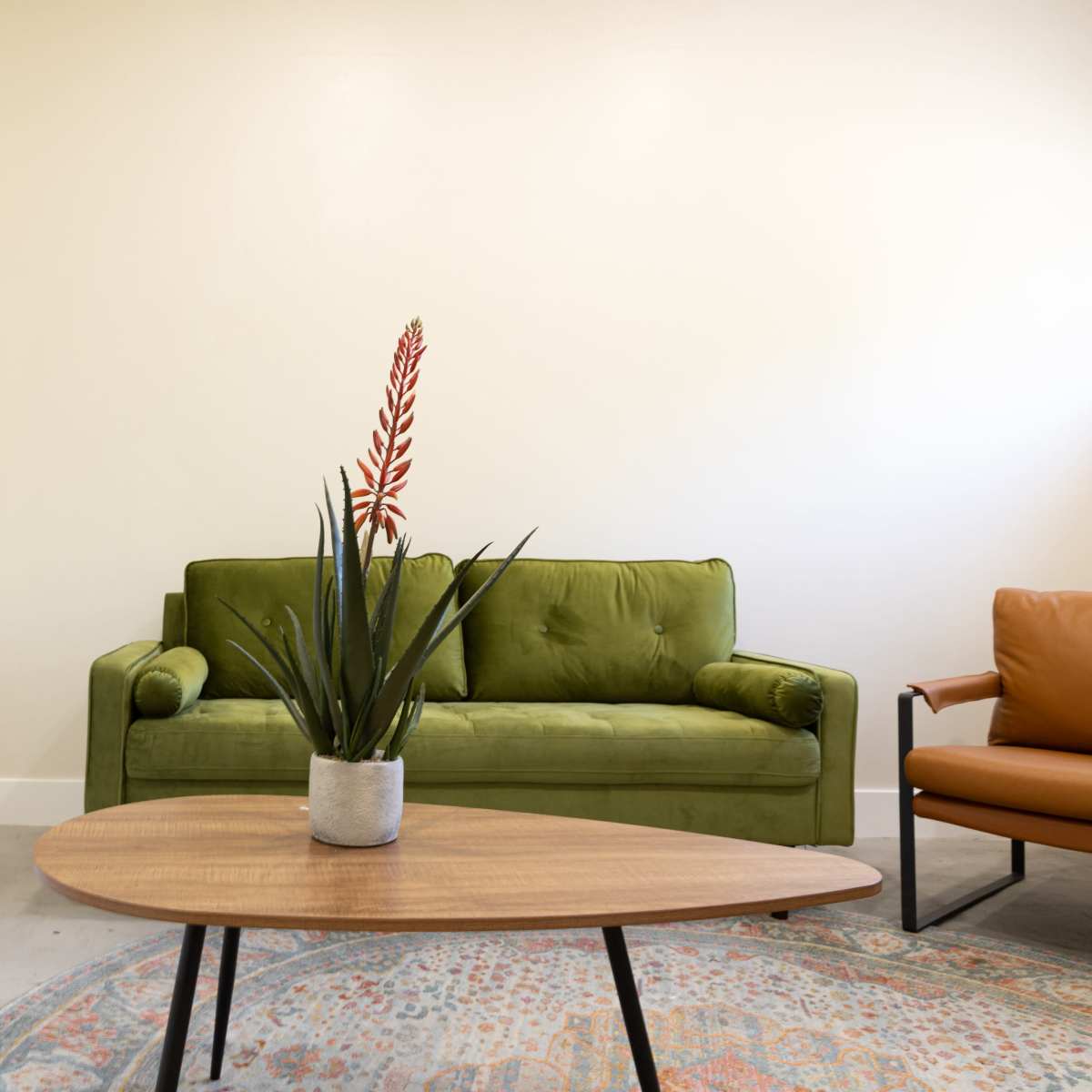 A green couch and an orange chair arranged around a wooden coffee table with a potted plant on it, set in a modern room with light-colored walls.