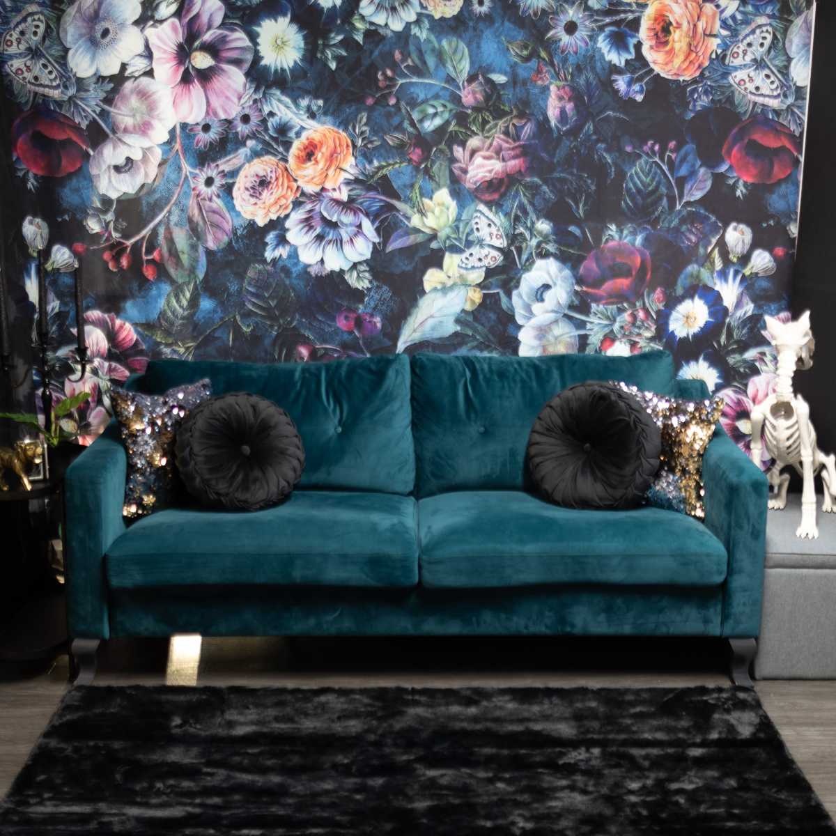 A teal sofa with black decorative pillows sits in front of a floral wallpaper and is accompanied by a decorative skeleton and a dark rug.