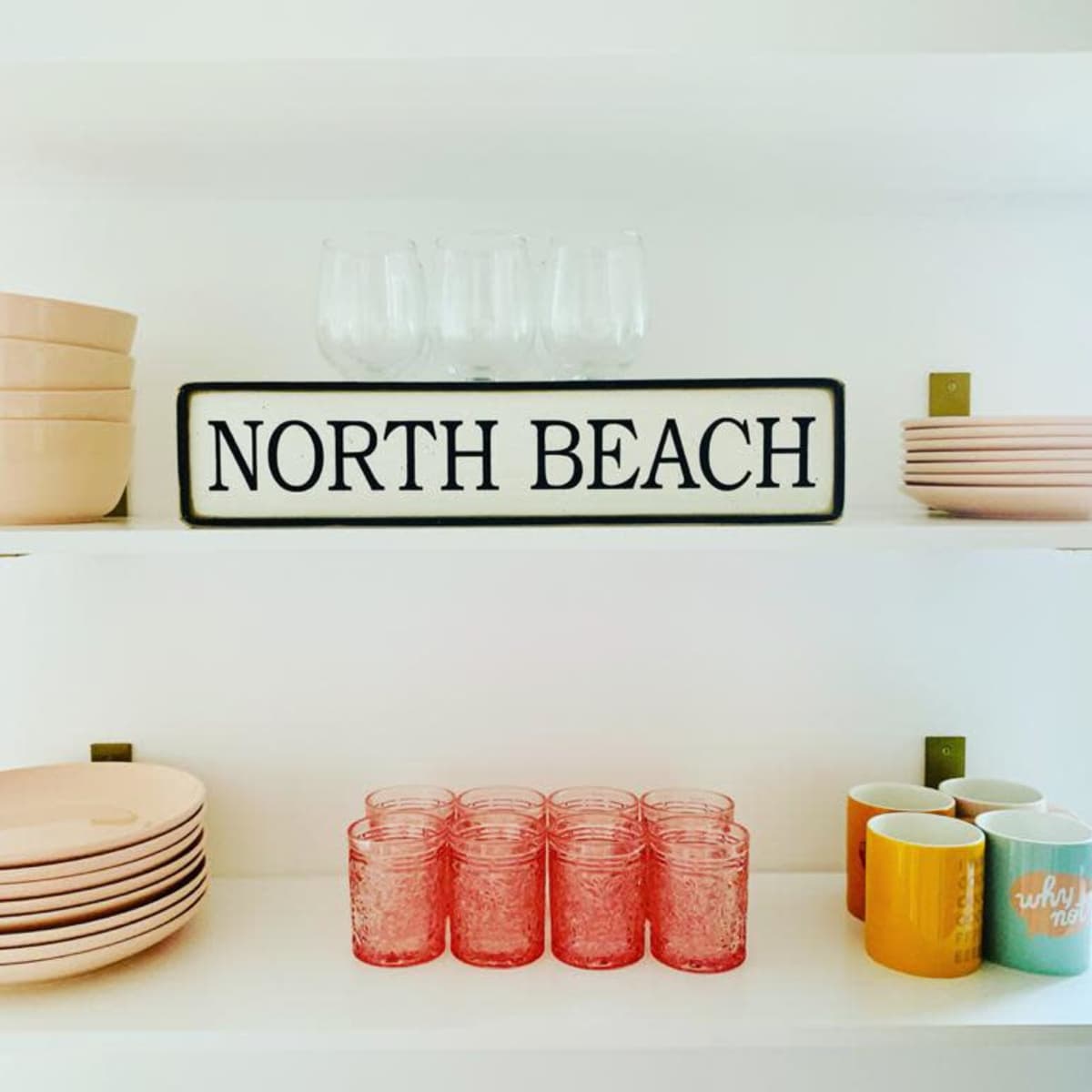 A white shelf displaying various kitchenware, including bowls, plates, cups, and glasses, along with a sign reading "NORTH BEACH."