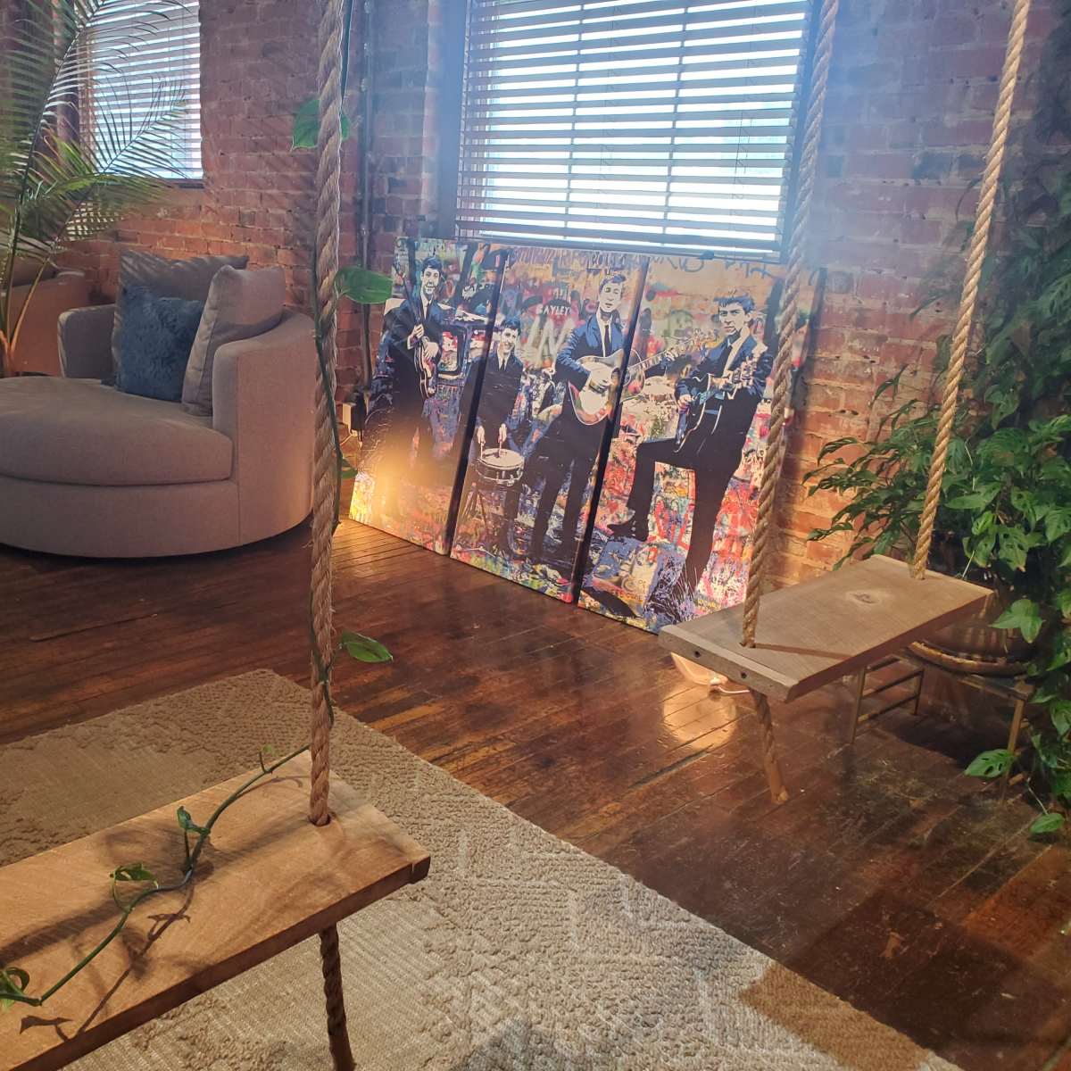 A room with a round gray sofa, a wooden swing, a large colorful painting of musicians, and plants positioned near brick walls and windows.