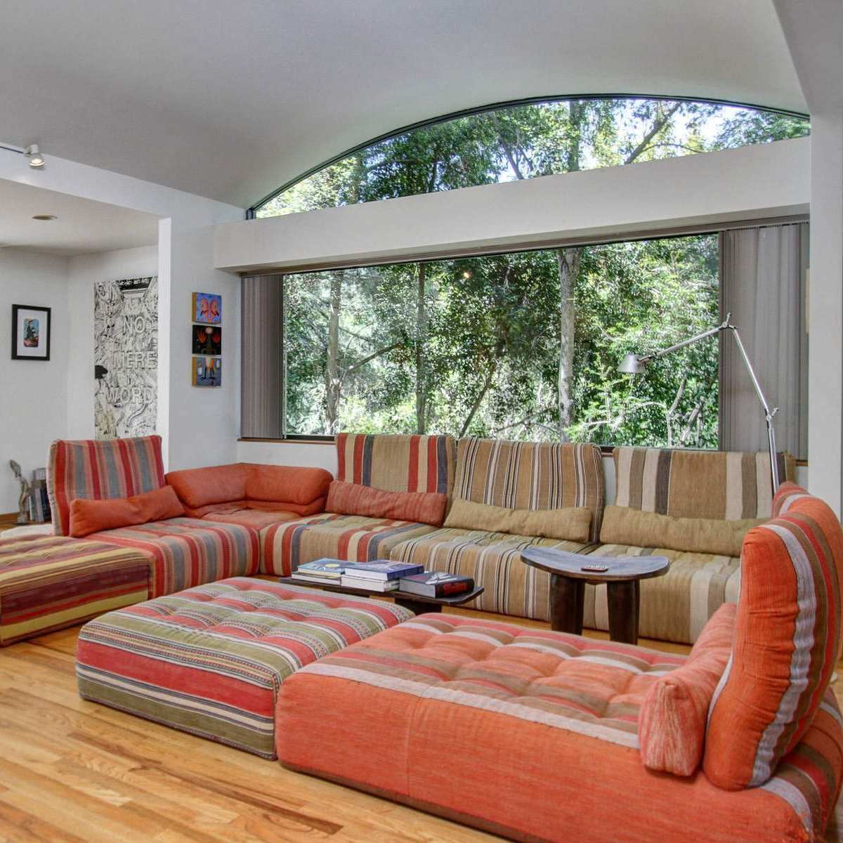 A spacious living room features a large, striped sectional sofa, wooden floors, and large windows overlooking greenery.