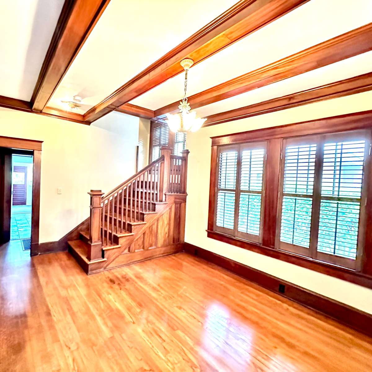 A room with wooden flooring, a staircase, and windows with shutters, all featuring wooden trim and a ceiling with decorative beams.