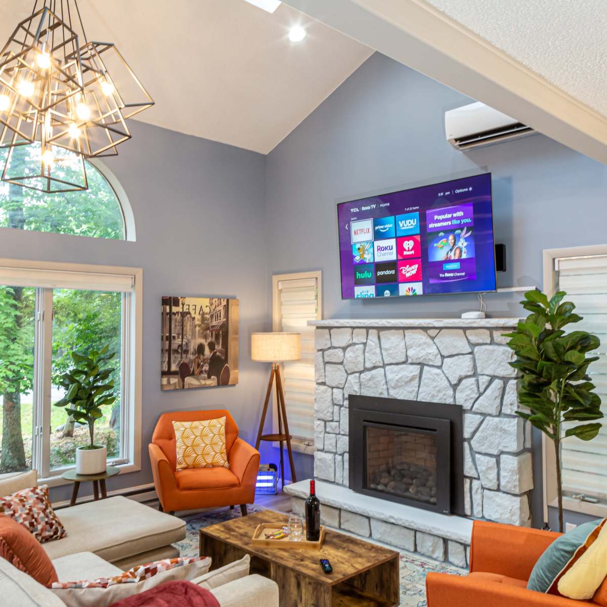 A modern living room features a stone fireplace, a large wall-mounted TV displaying streaming options, and comfortable seating arranged around a wooden coffee table.