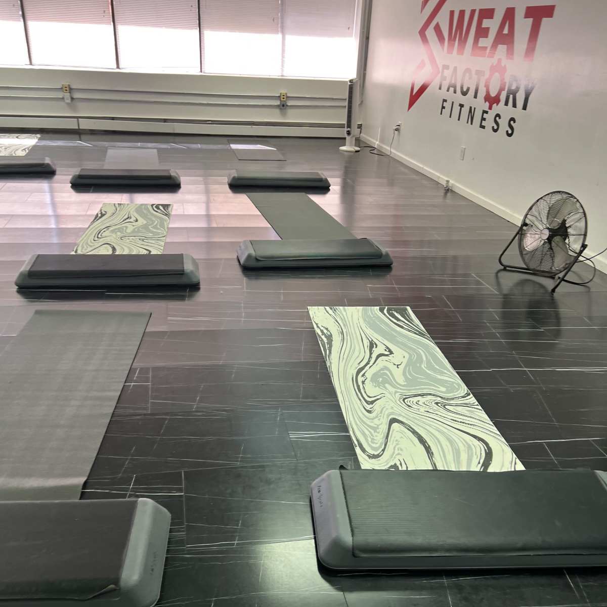 A fitness studio with several exercise mats arranged on a glossy black floor, along with a fan in the corner.