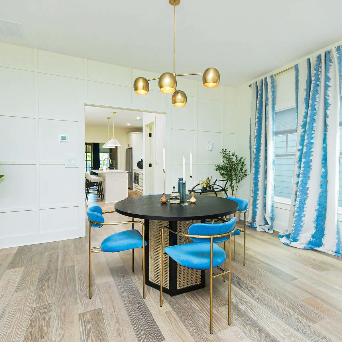 A dining area features a round black table surrounded by four blue chairs, with a background of patterned curtains and a doorway leading to another room.