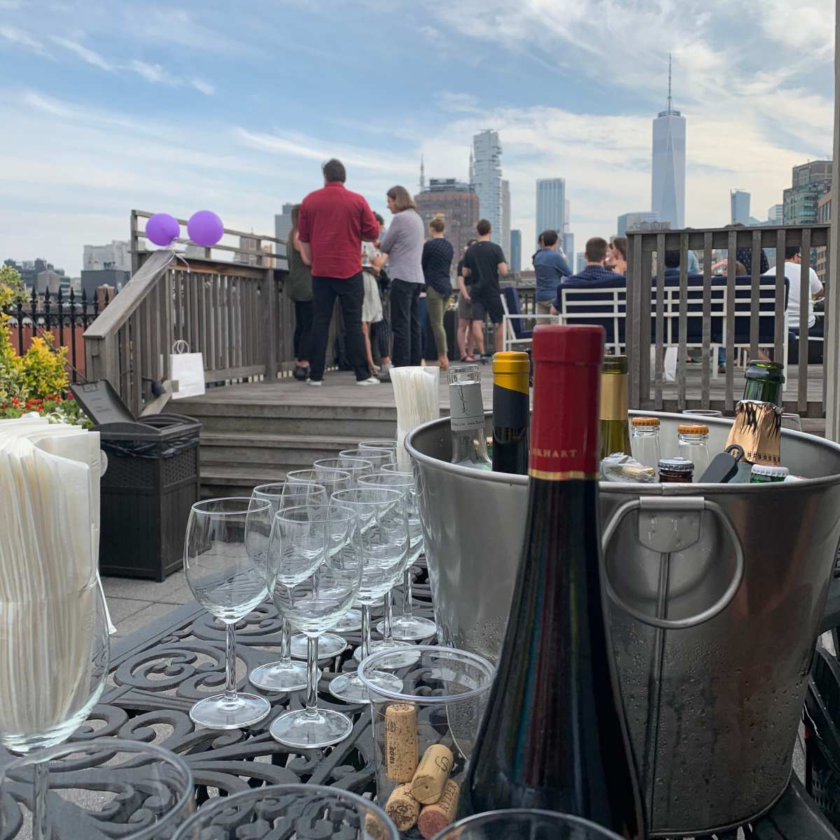 A rooftop gathering is taking place with people socializing in the background, while a table displays wine bottles and empty glasses in the foreground.