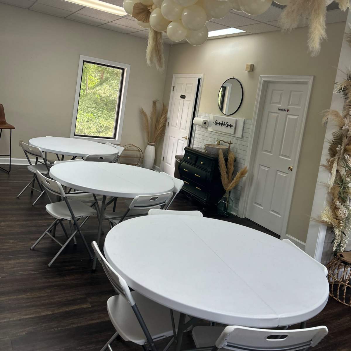 A lightly decorated event room with white circular tables, a faux fireplace, and a ceiling arrangement of pale balloons.