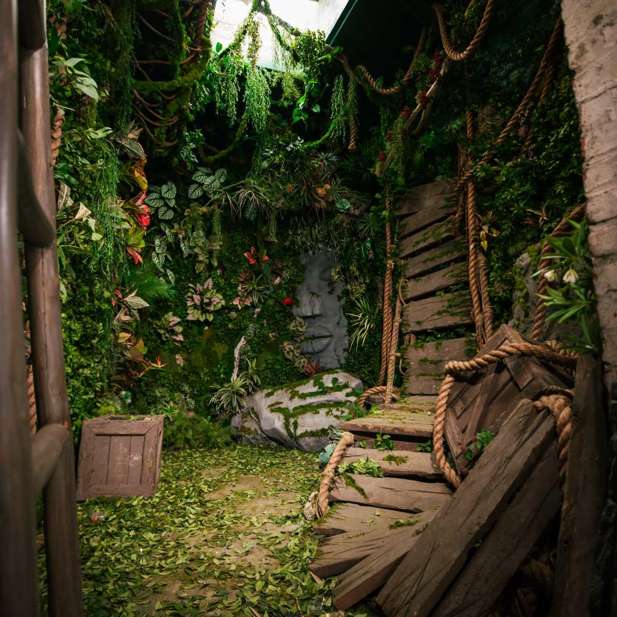 An indoor space overgrown with plants and vines, featuring a partially broken wooden swing and a crate amidst fallen leaves.