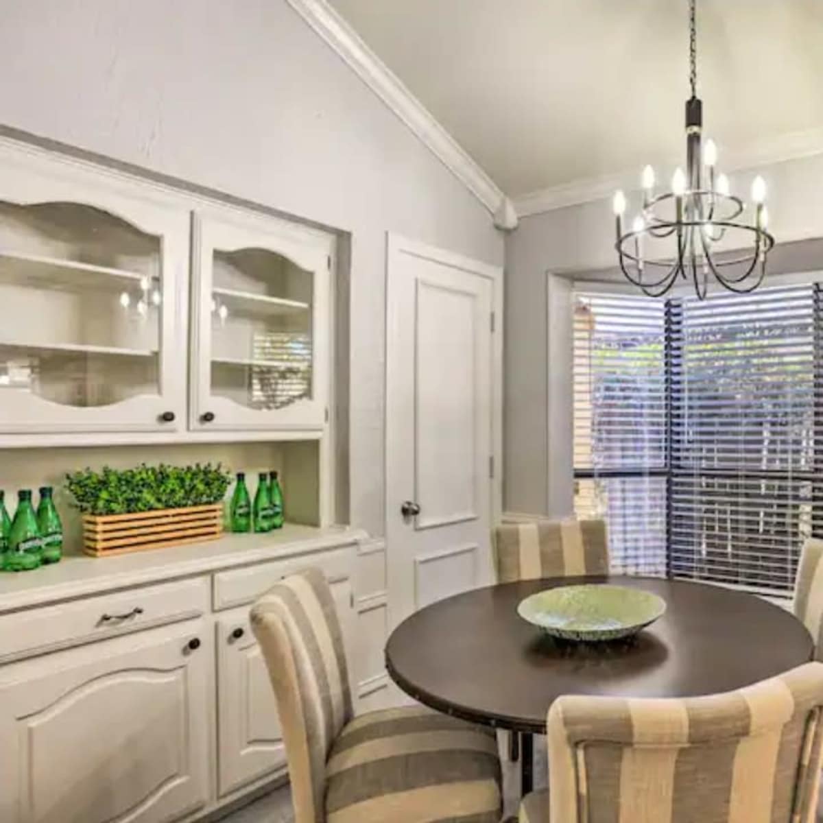 A dining area features a round table surrounded by four striped chairs, with a chandelier overhead and a window with blinds letting in light.