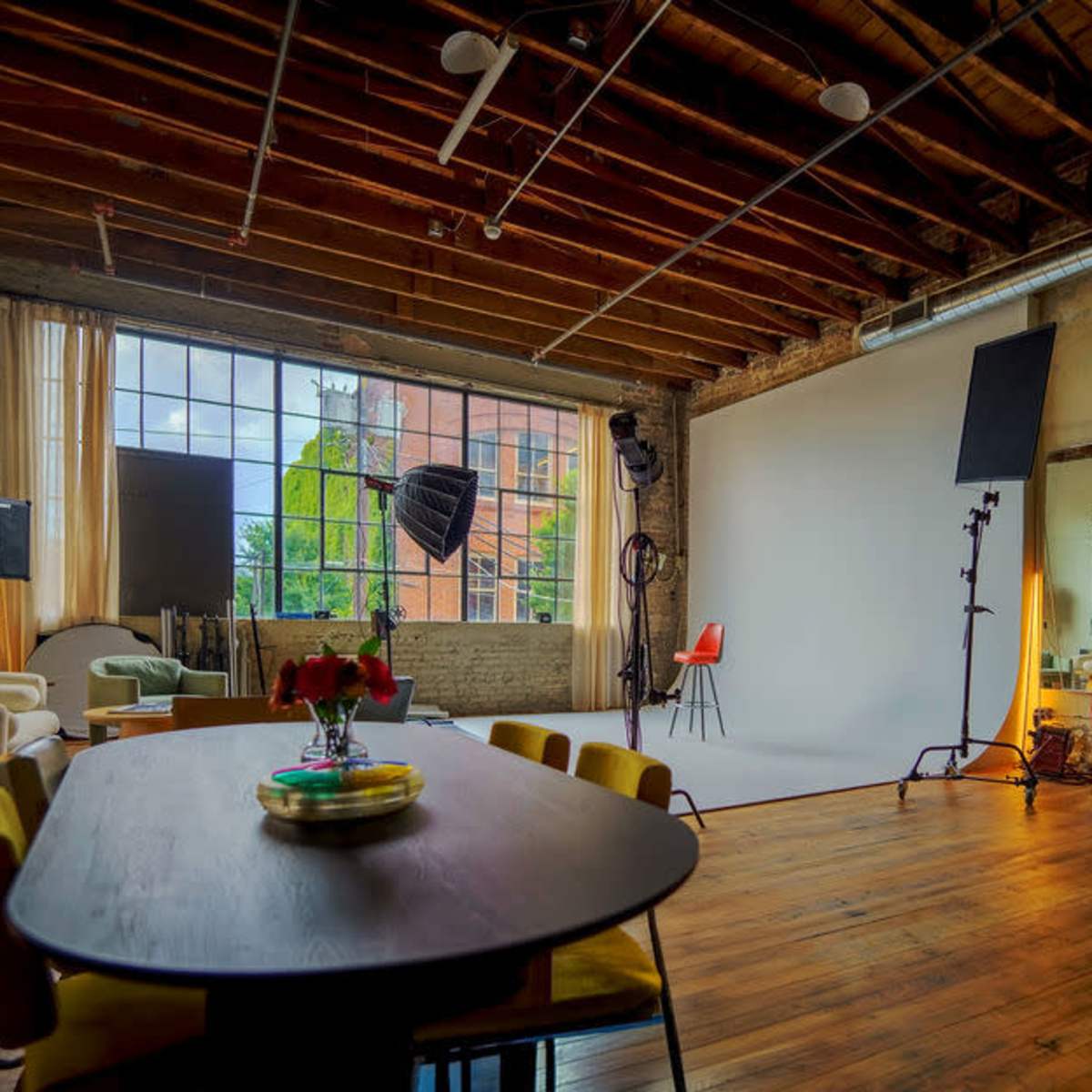 A spacious studio with wooden floors, large windows, and various furniture pieces, including a dining table and photography equipment.