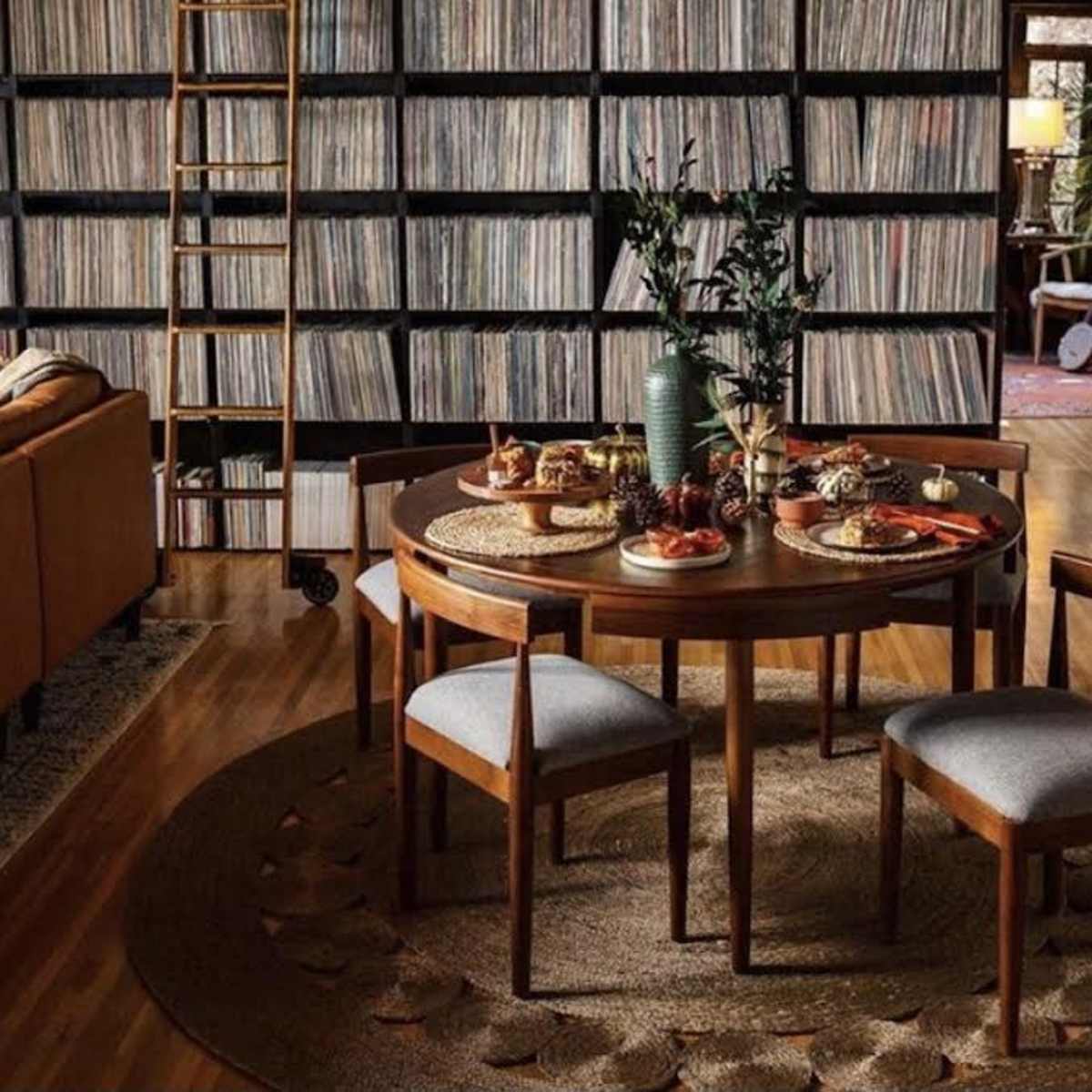 A round wooden dining table is set with food and surrounded by three chairs in a room featuring a large collection of vinyl records on shelves.