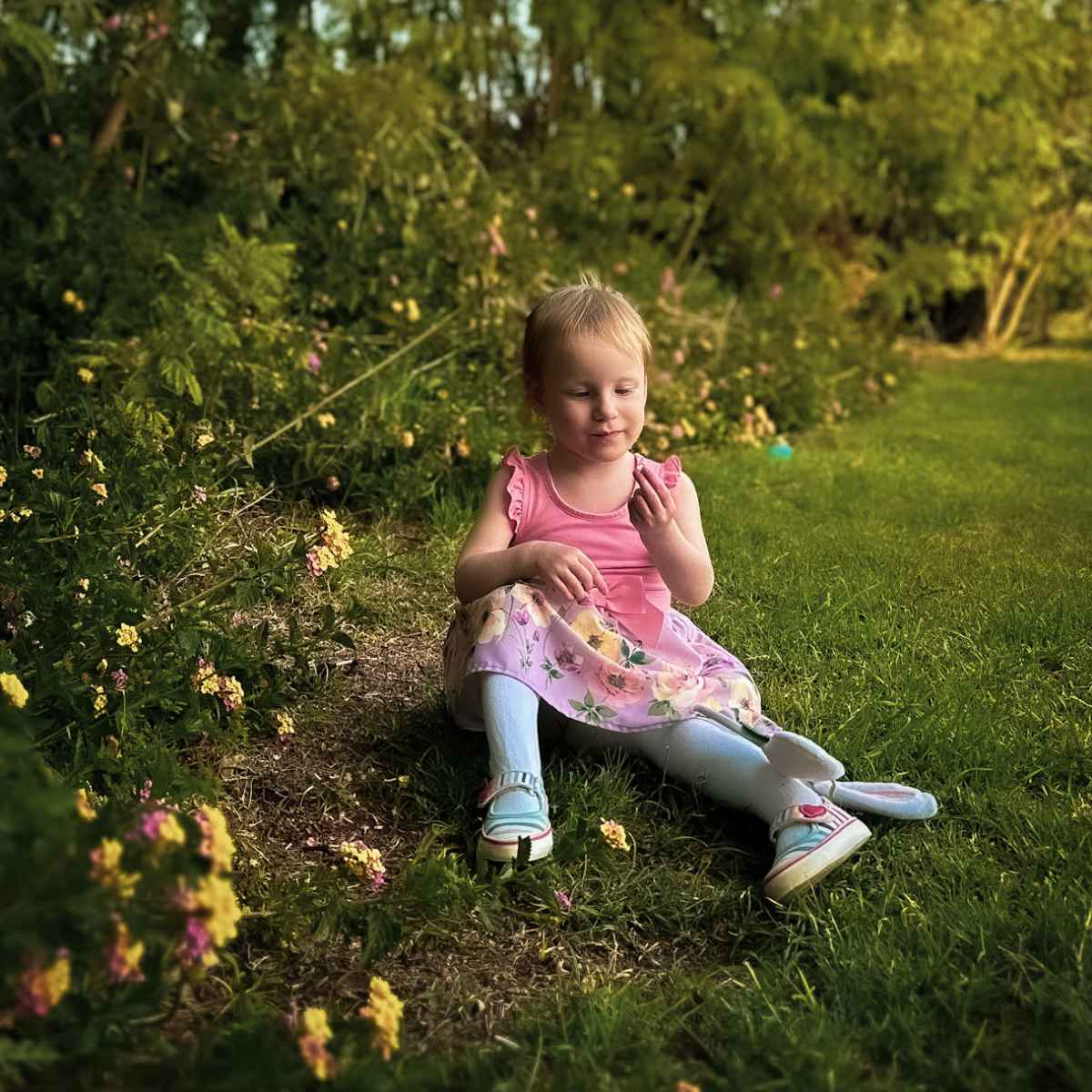 A child in a pink top and floral skirt sits on the grass, surrounded by greenery and flowers.