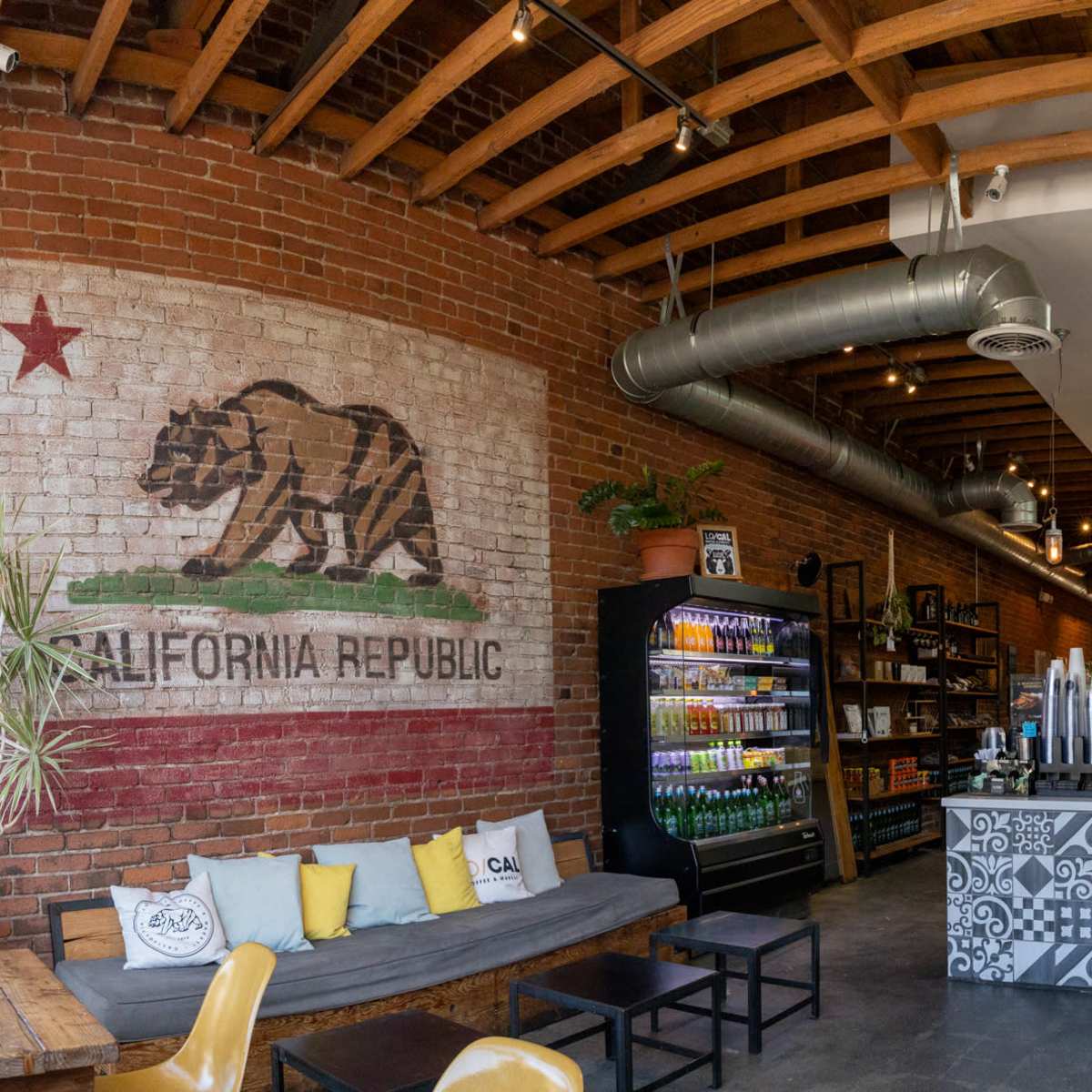 A café interior featuring a large mural of the California flag on a brick wall, a seating area with a long sofa, a refrigerator stocked with beverages, and a counter with coffee equipment.