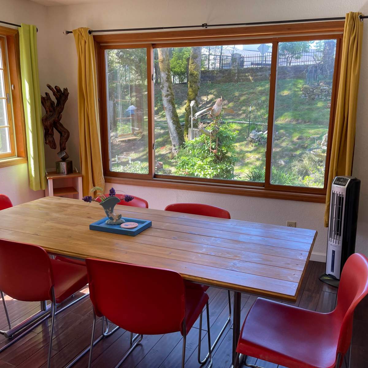 A wooden dining table with red chairs is positioned by a large window overlooking a green yard with trees.