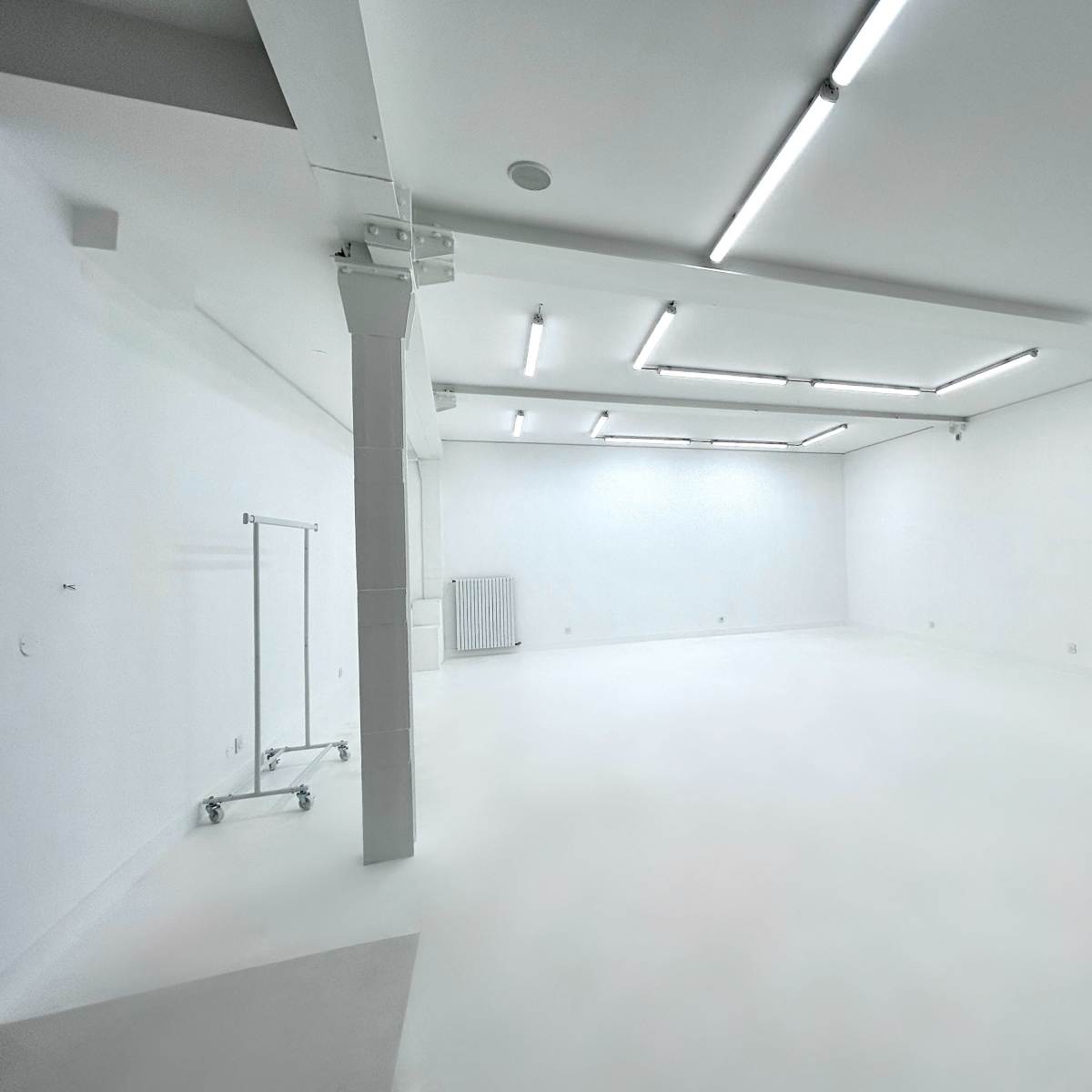 A spacious, minimalist room with white walls and a polished floor, featuring fluorescent ceiling lights and a clothing rack in one corner.