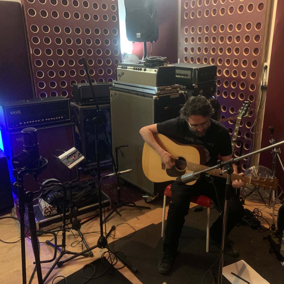 A musician sits on a chair playing an acoustic guitar in a recording studio surrounded by amplifiers and audio equipment.