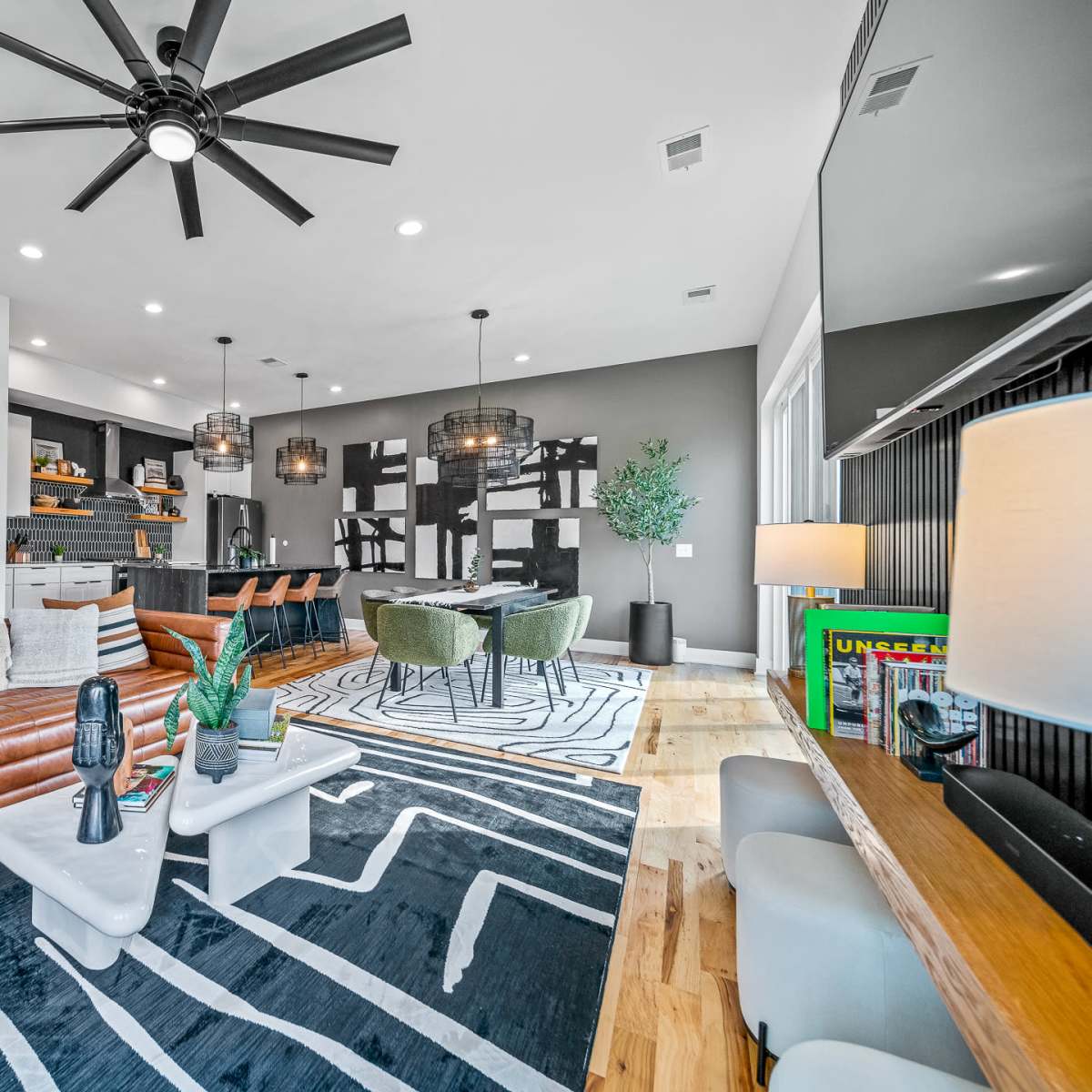A modern living room features a leather sofa, a striped area rug, and a kitchen area with dark cabinetry and pendant lighting.
