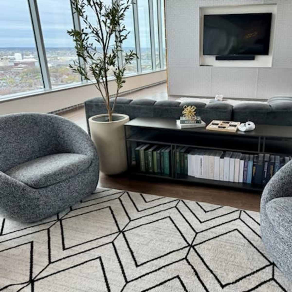 A modern living room features a gray sofa, two round chairs, a black coffee table with books, and large windows providing a city view.