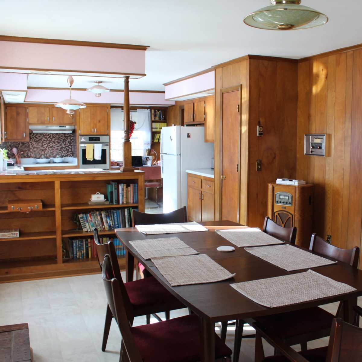 A wooden-furnished dining area with a table set for meals, adjacent to an open kitchen in a well-lit room.