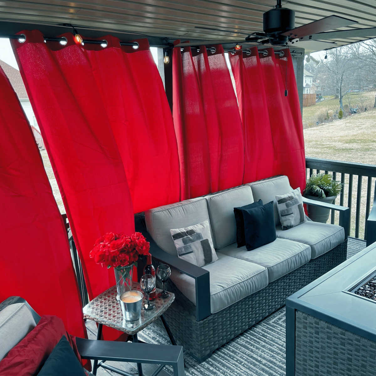 A covered porch features a gray seating area with red curtains, a table with a glass top, and decorative accents like a flower vase and candles.