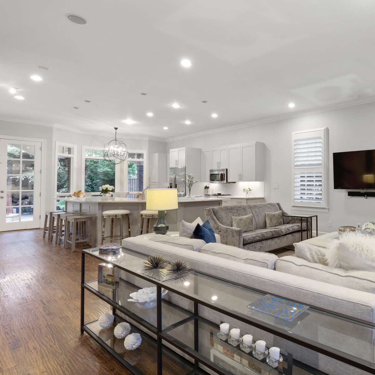 A modern living room with a light-colored sofa and a glass coffee table, adjacent to a kitchen area with white cabinetry and stainless steel appliances.