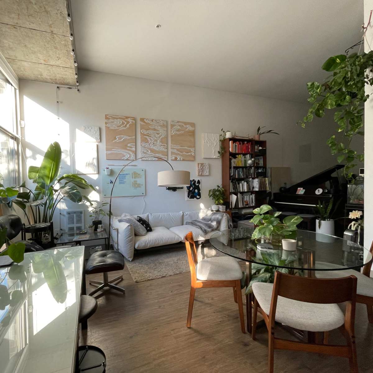 A bright and airy living space featuring a dining table, a sofa with decorative pillows, a piano, bookshelves, and various indoor plants.