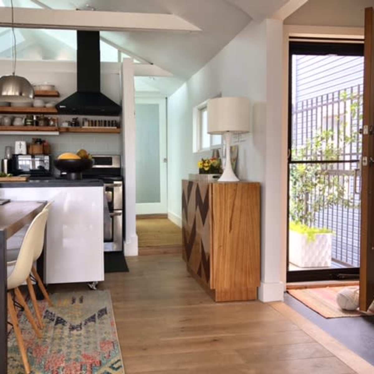 A modern kitchen and dining area with a wooden dining table, open shelving, and a front door leading to an exterior space.