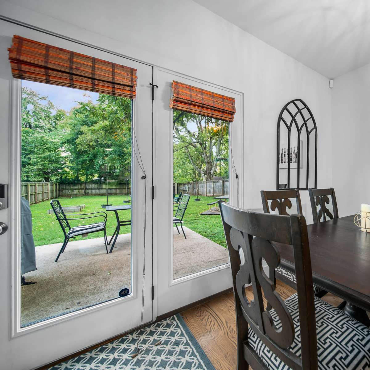 A dining area features a dark wooden table surrounded by chairs, with glass doors that open to a backyard visible through the open entrance.