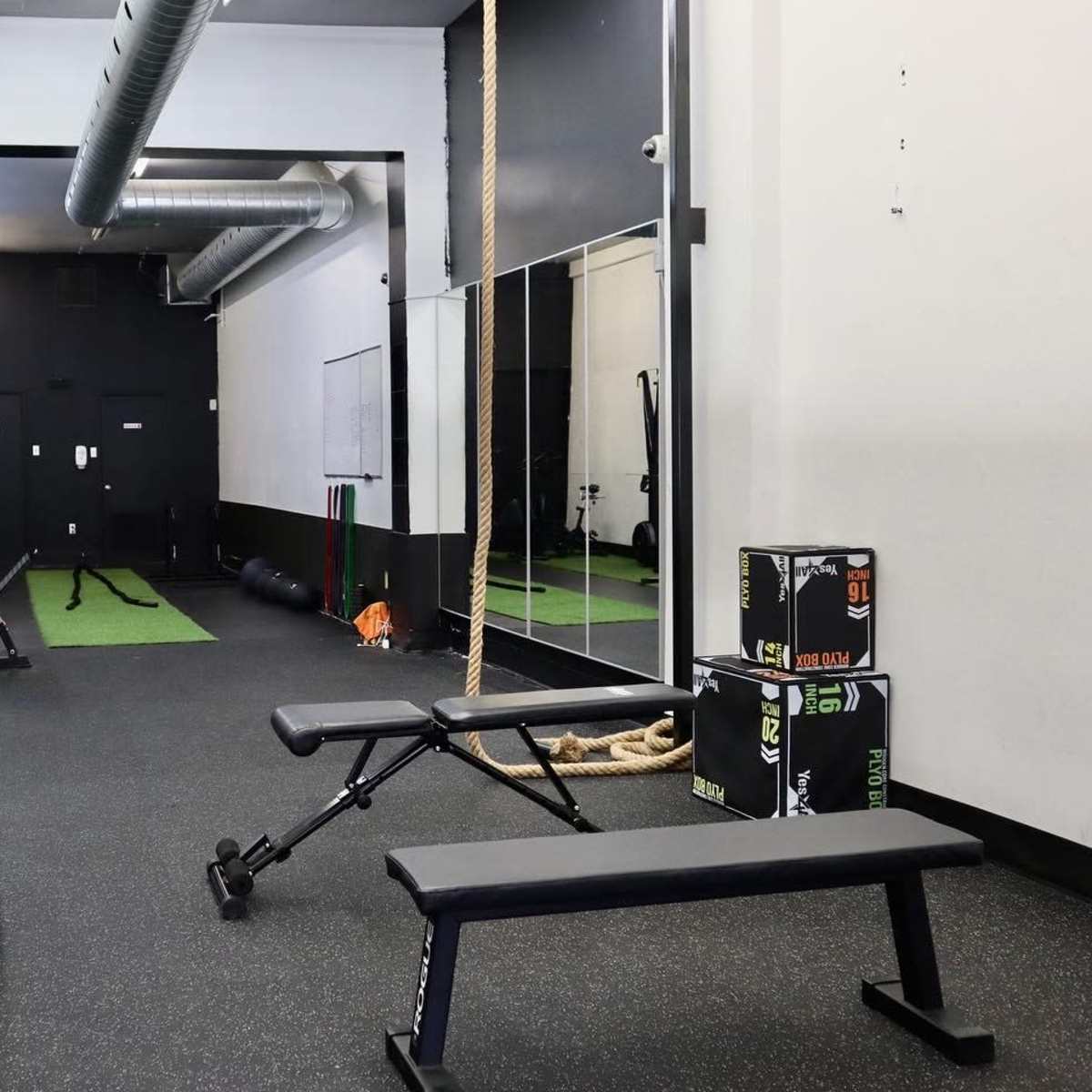 A fitness gym area featuring dumbbells, exercise benches, and a mirrored wall alongside gym equipment and a climbing rope.