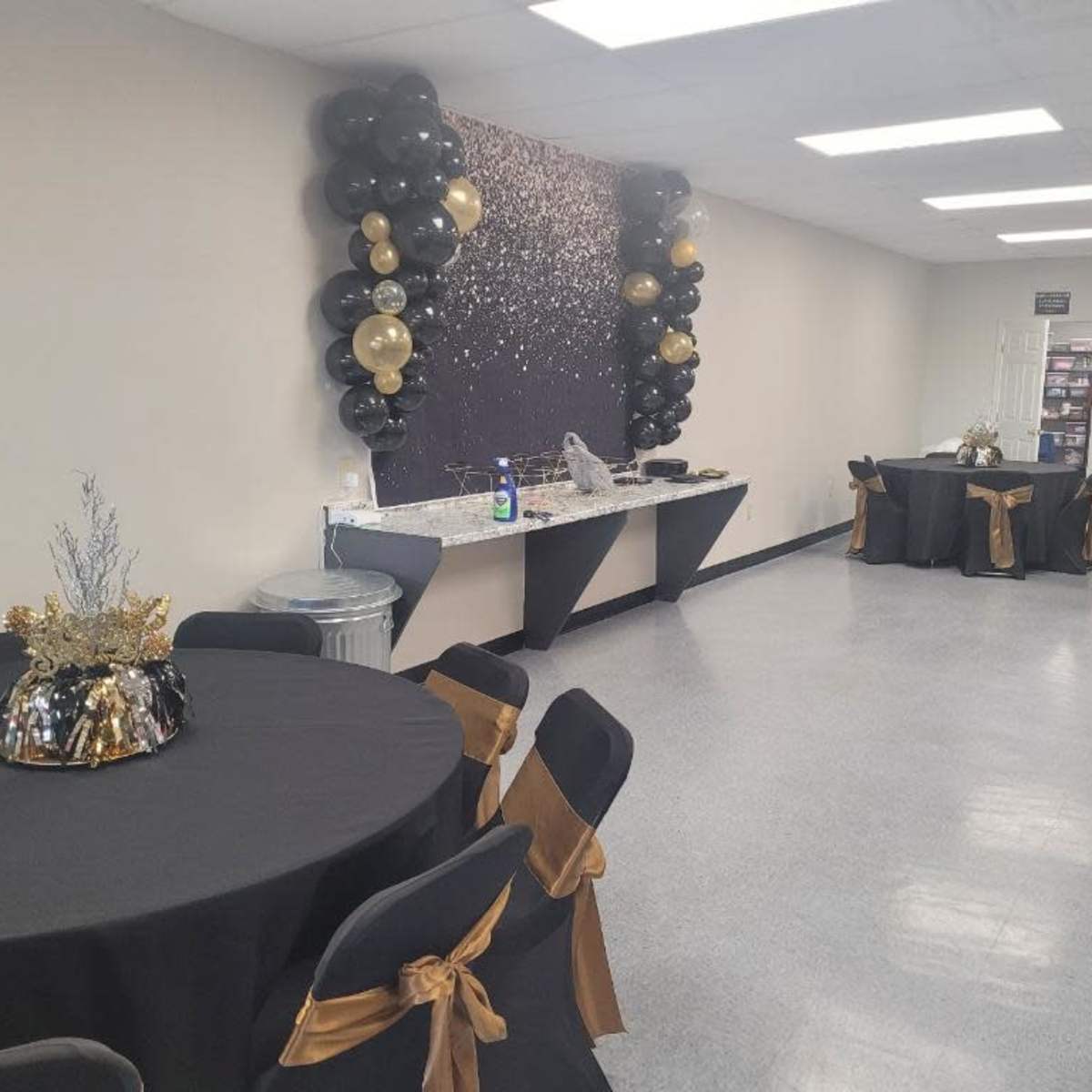 A decorated event space featuring black tablecloths with gold accents, a balloon backdrop, and a food preparation area along one wall.