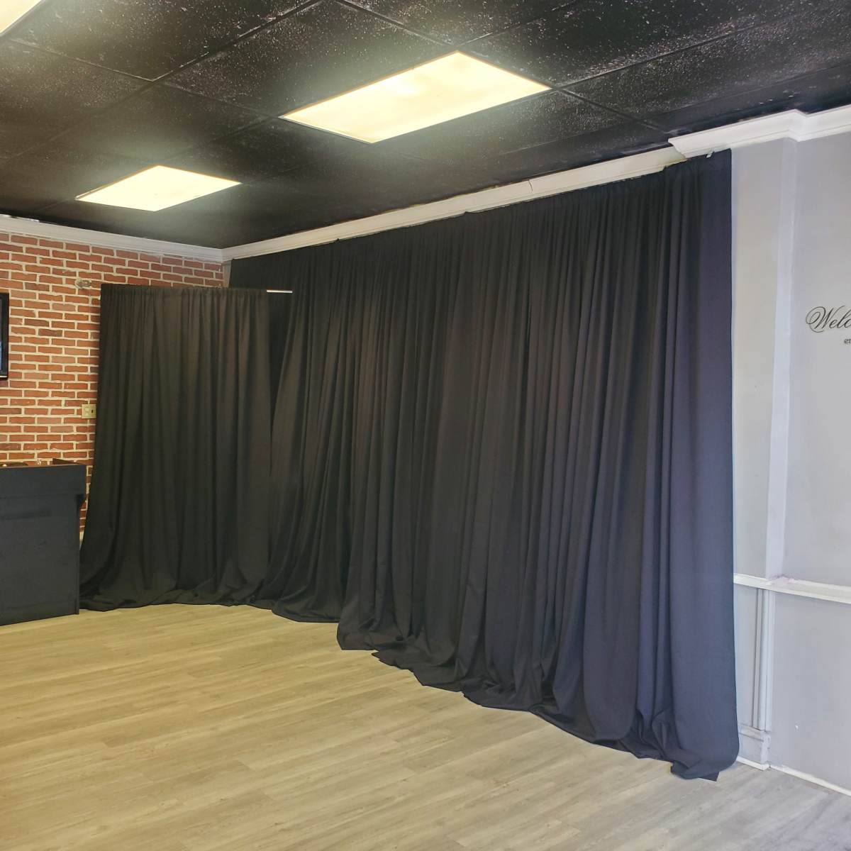 A room with black curtains draped along two walls, a brick accent wall, and a light-colored wooden floor.