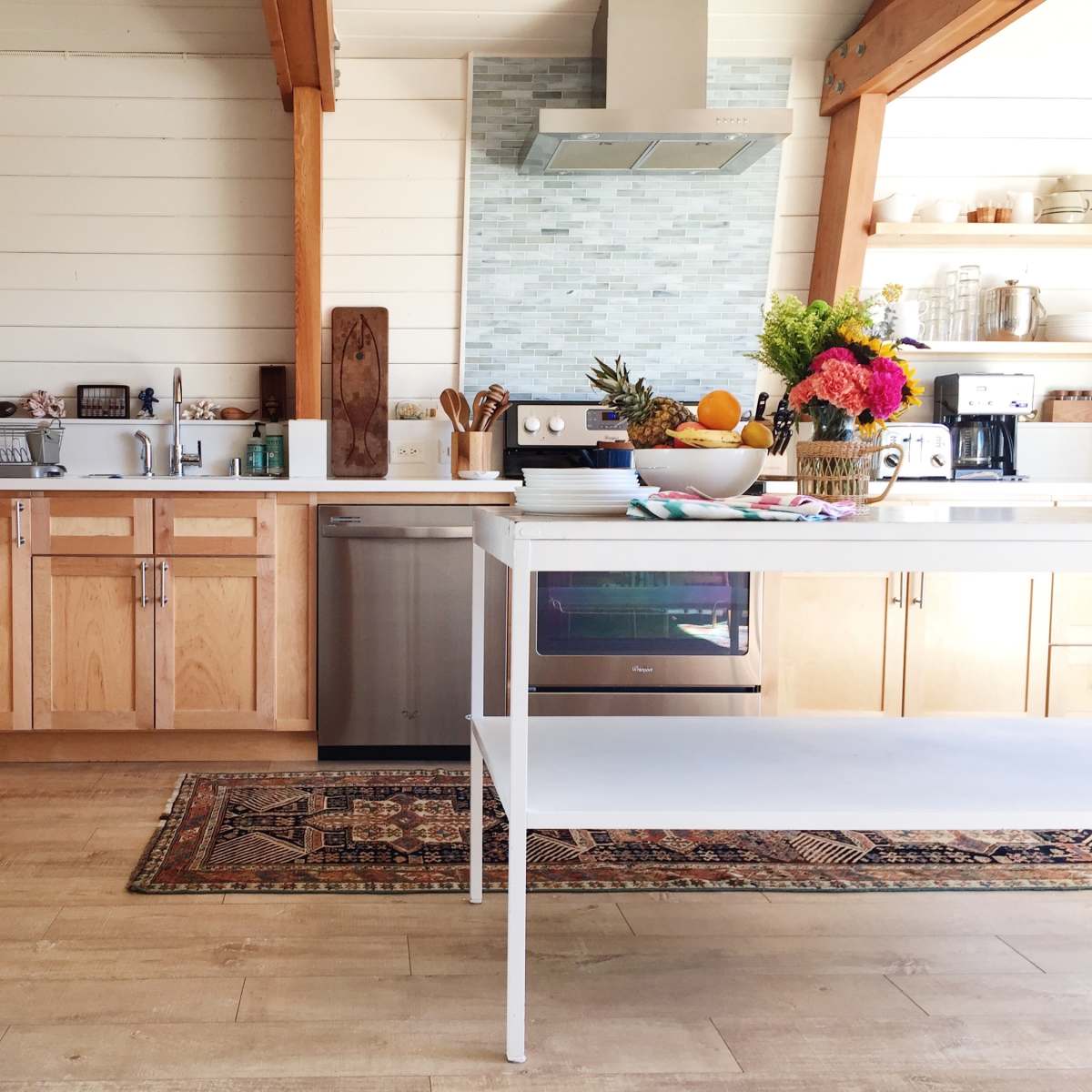 A modern kitchen features a stainless steel refrigerator, wood cabinetry, a bright countertop with a fruit arrangement, and a decorative rug on the floor.