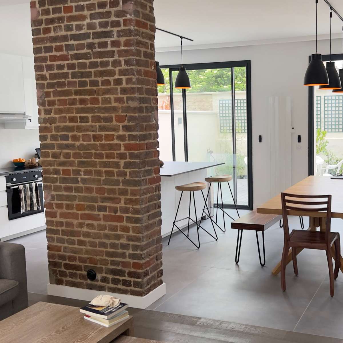 A modern open-plan living space with a brick column, a dining area with wooden furniture, and a kitchen visible in the background.