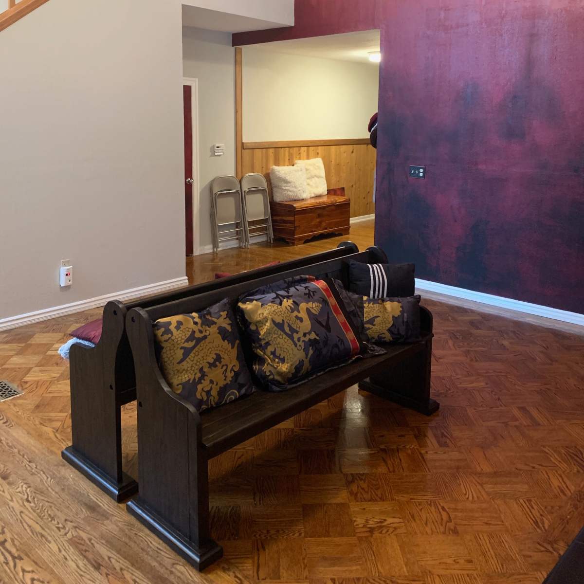 A living room with a dark red accent wall, a wooden bench adorned with patterned cushions, and a staircase in the background.