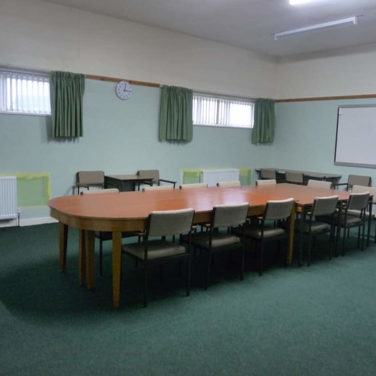 A large, oval table surrounded by chairs is set in an empty meeting room with green carpeting and small windows.