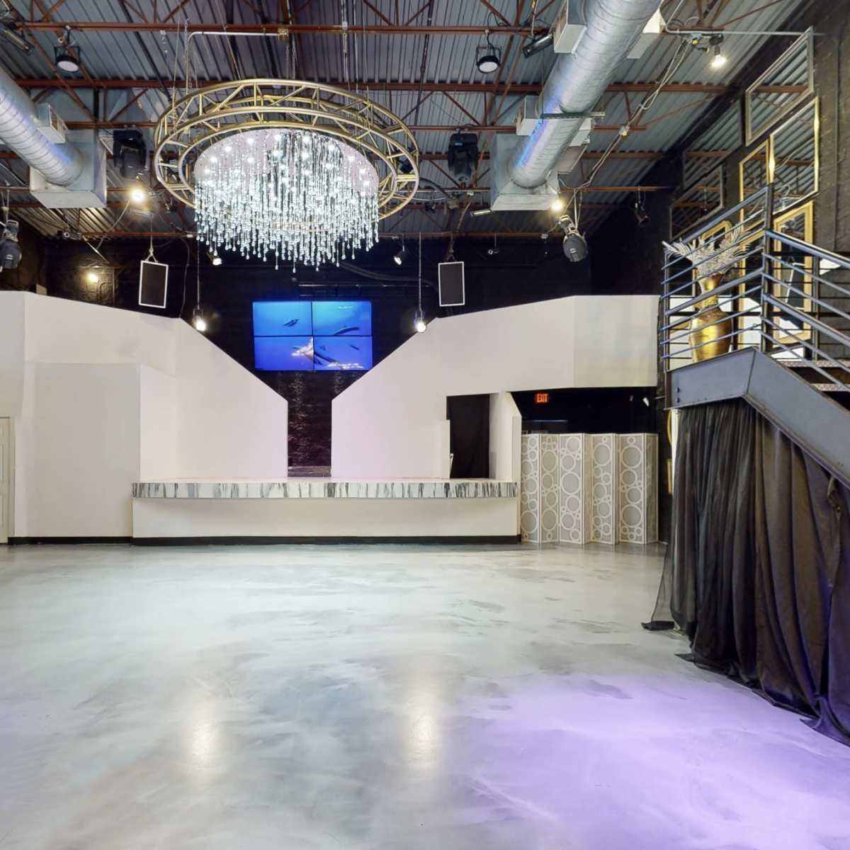 A spacious event venue with a central chandelier, a bar area at the back, and two staircases leading to an upper level.
