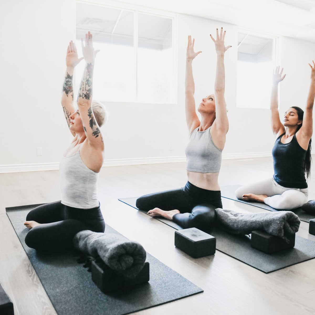 Three women practice yoga in a bright studio, each stretching with their arms raised while seated on mats.