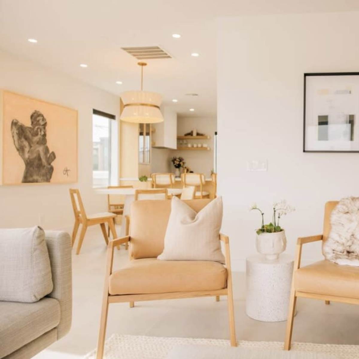 A modern living room with two light brown armchairs, a small side table, and a minimalist decor featuring a wall art piece and a dining area in the background.
