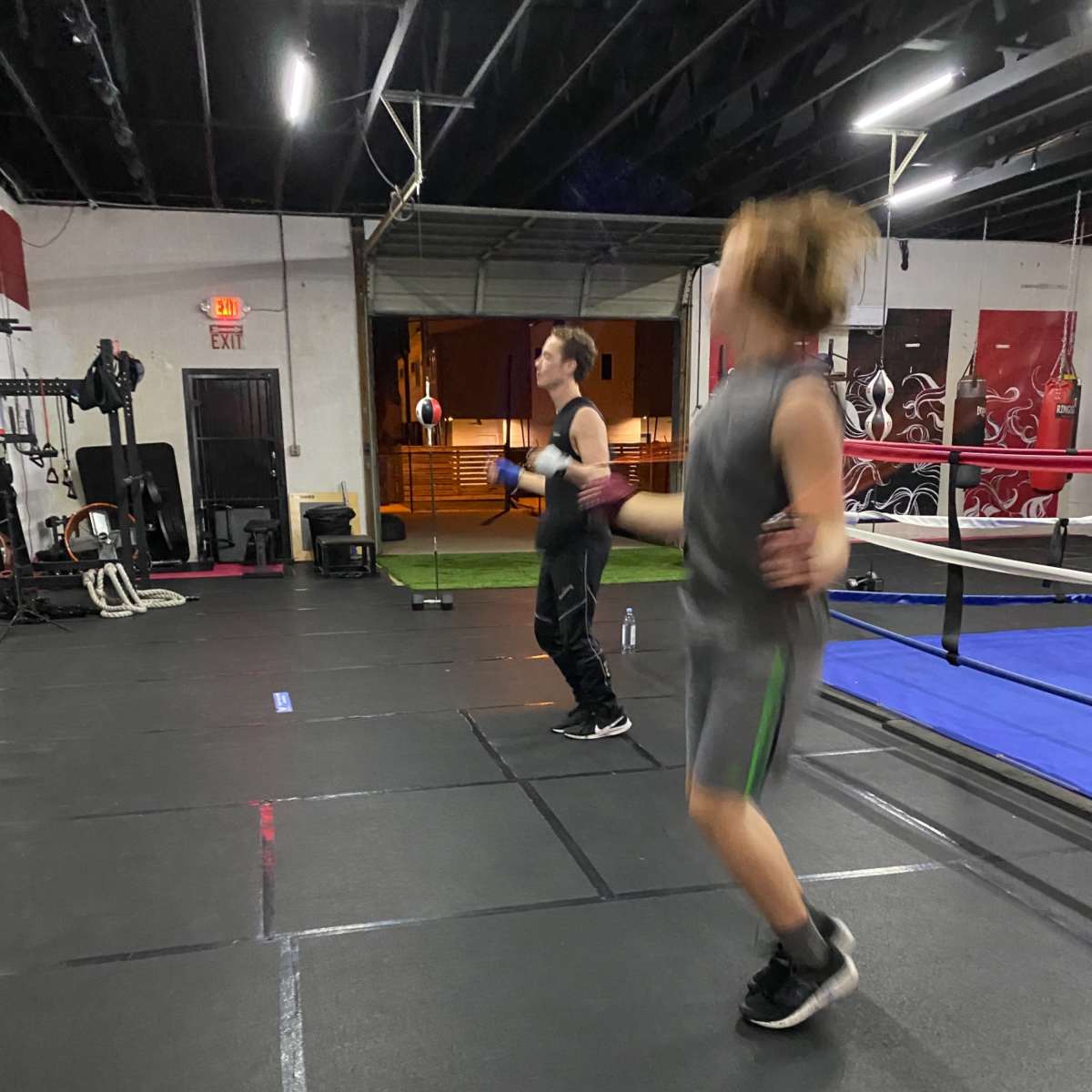 Two individuals are jumping rope in a gym with workout equipment and a boxing ring in the background.