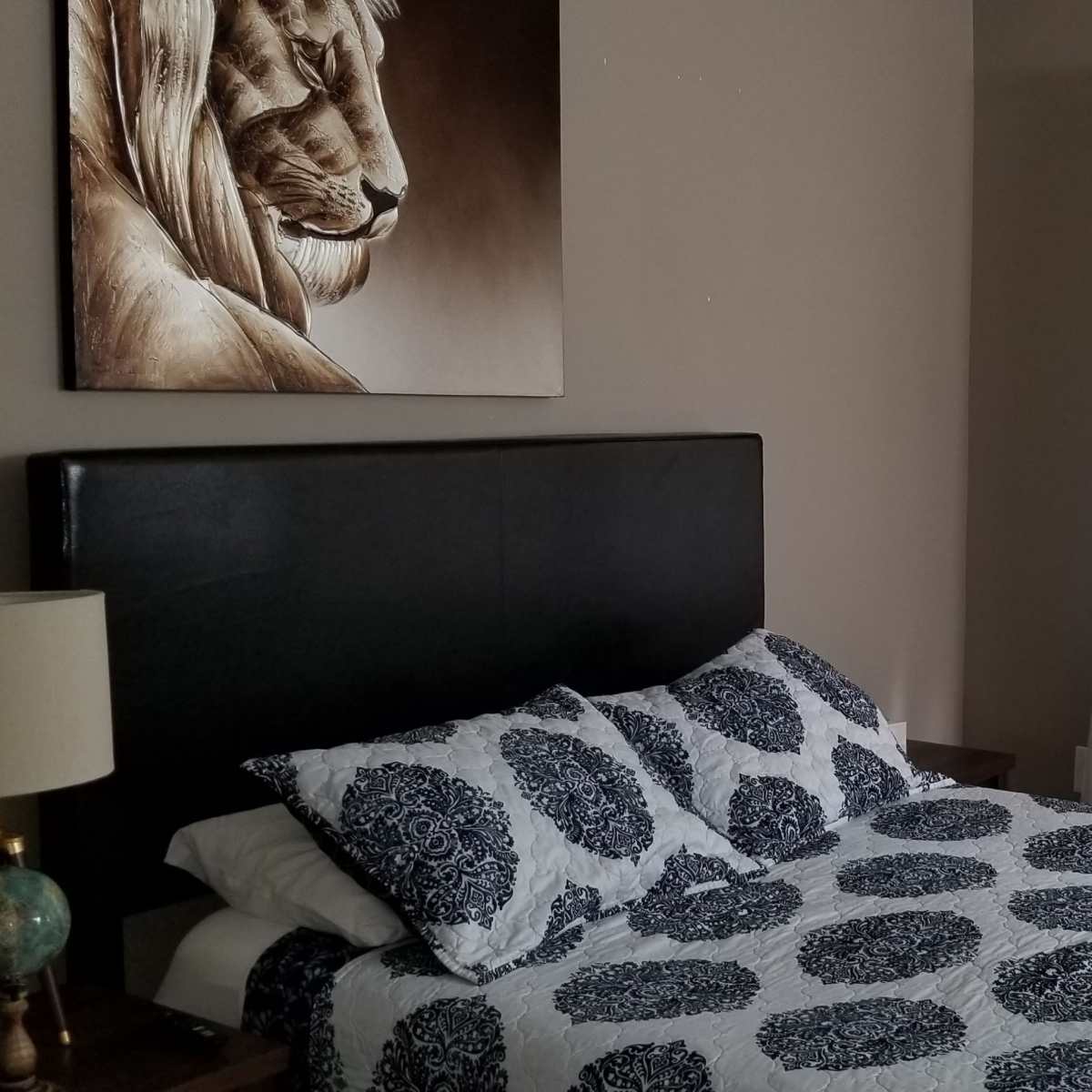 A bedroom features a bed with a patterned blue and white bedspread, a lion portrait on the wall, and sheer white curtains allowing light to enter.