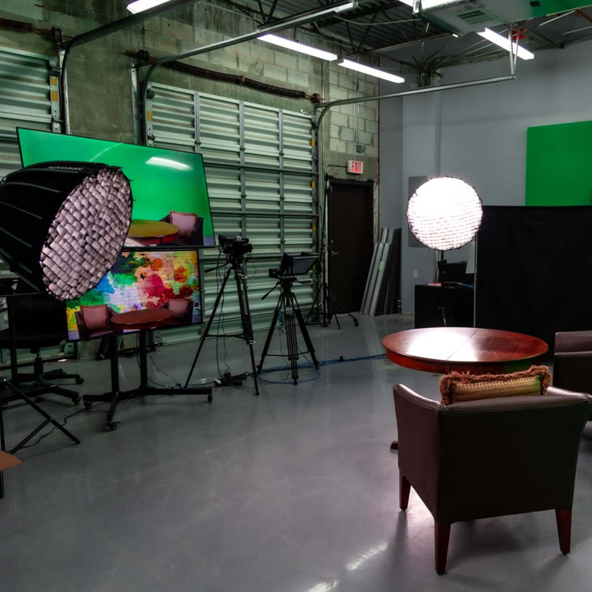 A video production studio with two chairs positioned near a round table, surrounded by lighting equipment and a green backdrop.