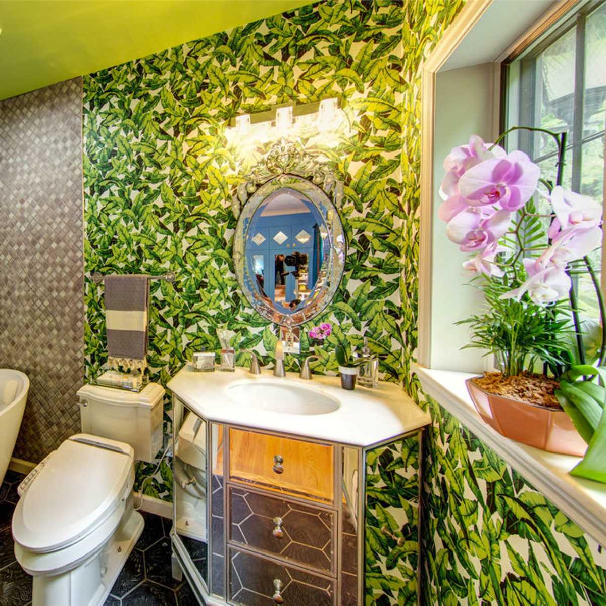 A small bathroom featuring a freestanding bathtub, a modern vanity with a mirror, and leafy wallpaper accented by a decorative light fixture.