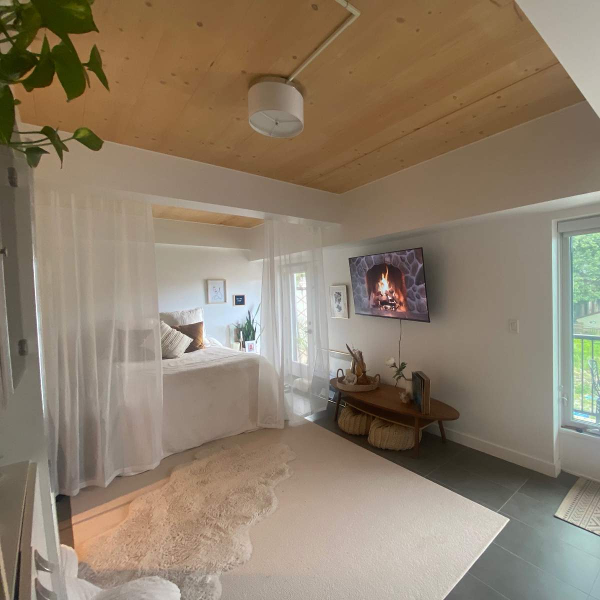 A cozy room features a bed draped with sheer curtains, a wall-mounted television displaying a fireplace, and a balcony door leading to an outdoor view.