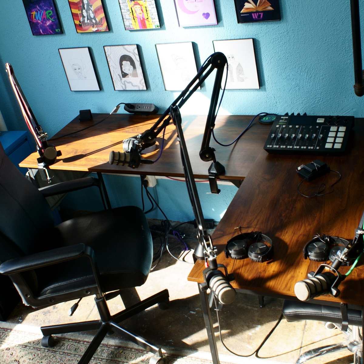 A bright blue room featuring two desks with microphones, headphones, and a control panel, surrounded by framed artwork on the walls.