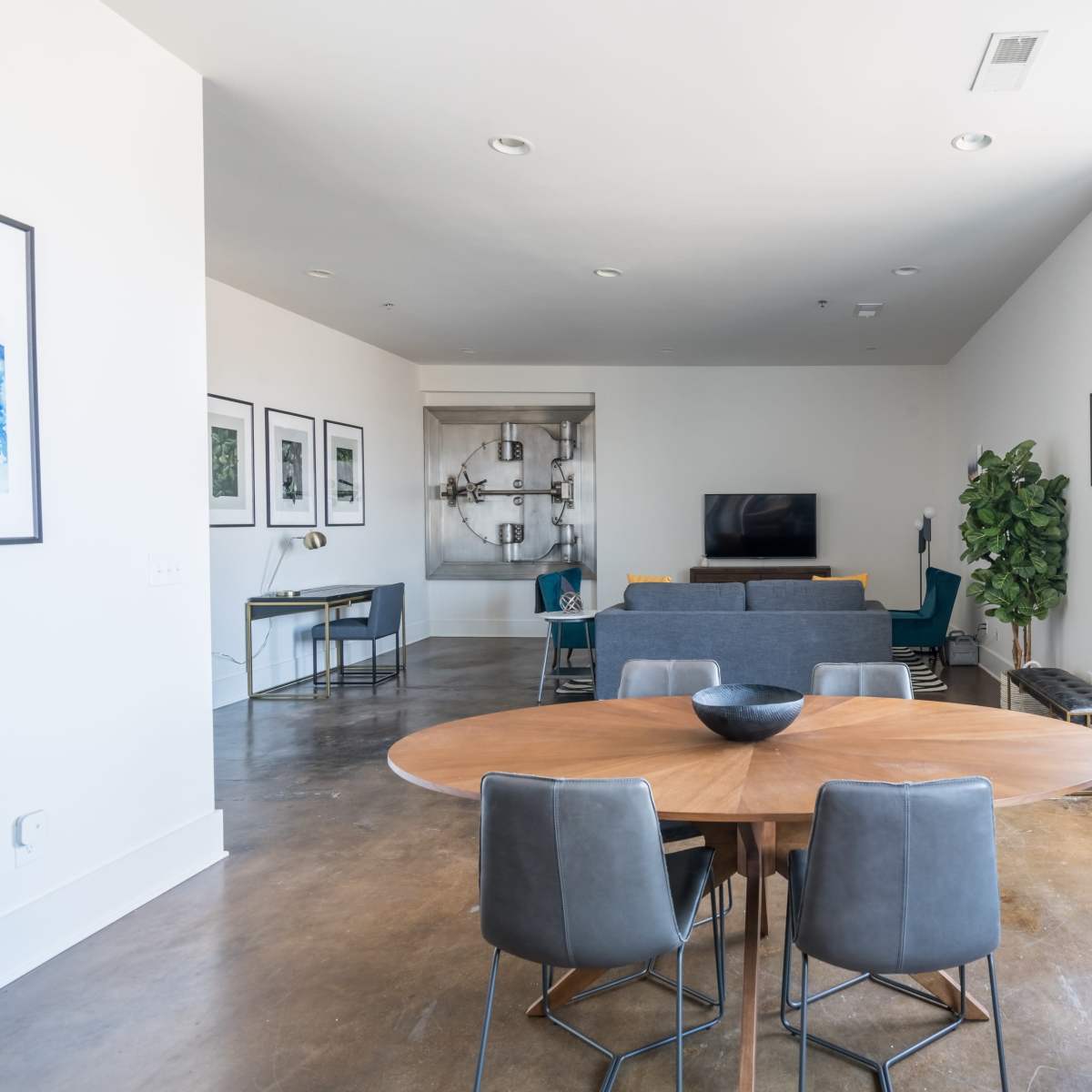 A modern living space with a dining area featuring a round wooden table surrounded by gray chairs, and a living room with a couch and television in the background.