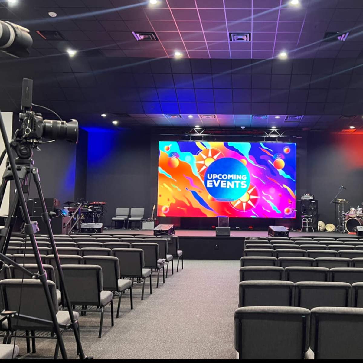 An auditorium set up for an event, featuring a large screen displaying "UPCOMING EVENTS" and rows of chairs facing the stage.
