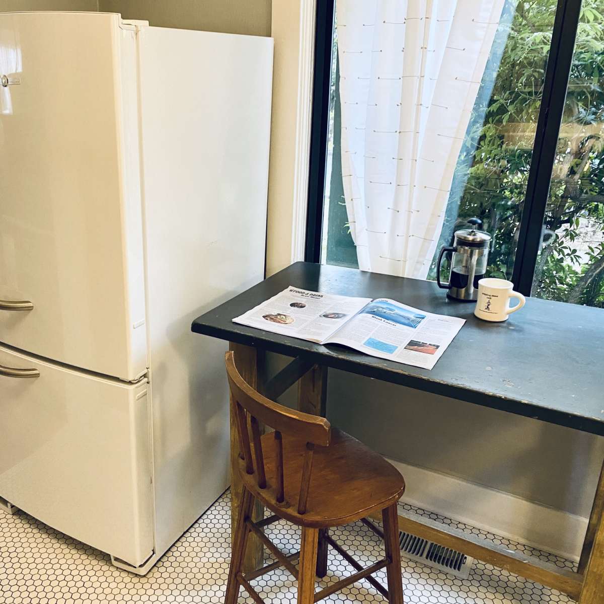 A white refrigerator stands next to a small dining table with a magazine and a coffee cup on it, set against a backdrop of a window and greenery outside.
