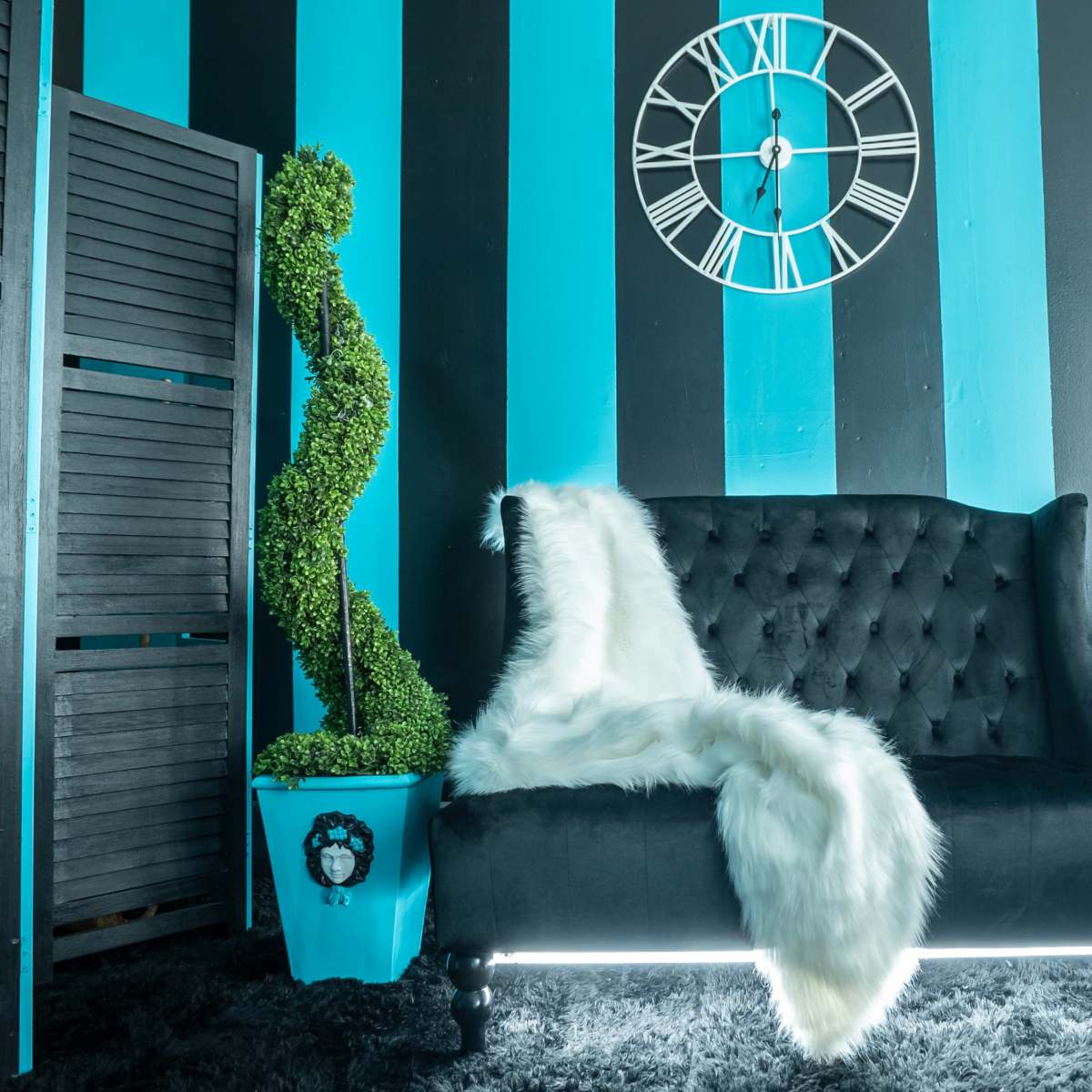 A black tufted sofa adorned with a white fur throw, positioned against blue and black striped walls, accompanied by a clock, a decorative plant, and a glass side table with a vintage telephone.